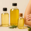 Φυσικά λάδια σώματος σε γυάλινα μπουκάλια με εκχυλίσματα όπως καρύδα, αμύγδαλο και ελιά, δίπλα σε γυναικείο χέρι που εφαρμόζει λάδι στο δέρμα.